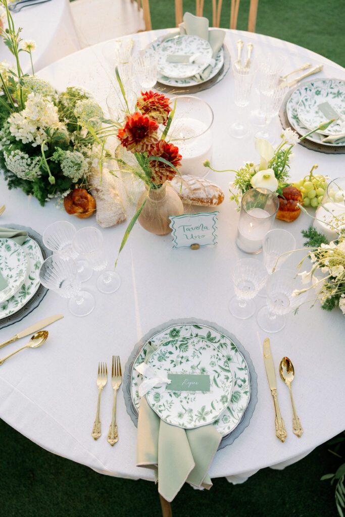 Elegant autumn dinner tablescape featuring green floral-patterned plates, gold flatware, and crystal glassware on a white linen tablecloth.